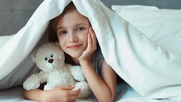 Meisje knuffelen teddybeer onder de deken en glimlachend in de camera. Zoomen — Stockvideo