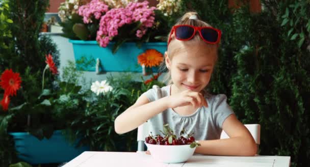 Lány eszik a cseresznye. Nagy szemekkel. Gyermek étkezési bogyók, és csodálja és látszó-on fényképezőgép. Hüvelykujj-fel. oké — Stock videók