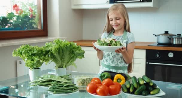 Девушка держит миску салата и предлагает зрителям. Молодой шеф-повар с овощами возле кухонного стола и улыбающийся в камеру. Девушка пропагандирует здоровое питание — стоковое видео