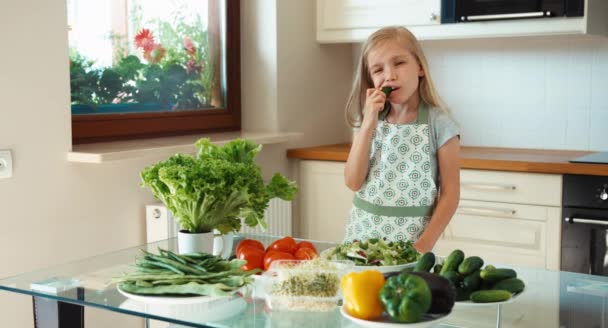 キュウリを食べるとカメラに笑顔の女の子の若いシェフ。親指を。わかりました — ストック動画