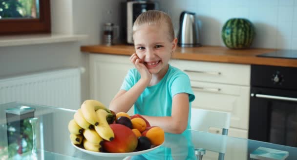 La ragazza fa pubblicità alla frutta. Bambino seduto sul tavolo della cucina e ridendo della macchina fotografica. Pollice in alto. Va bene. — Video Stock