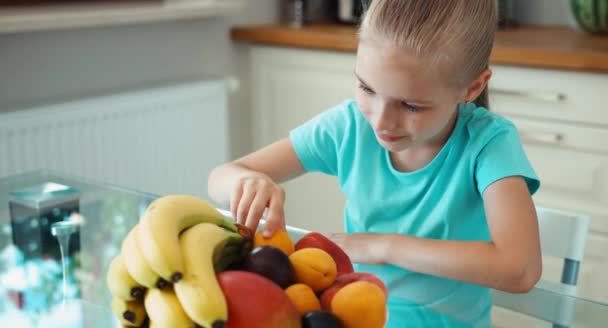 Девушка ест абрикос. Ребенок сидит на кухонном столе рядом с большими тарелками фруктов — стоковое видео