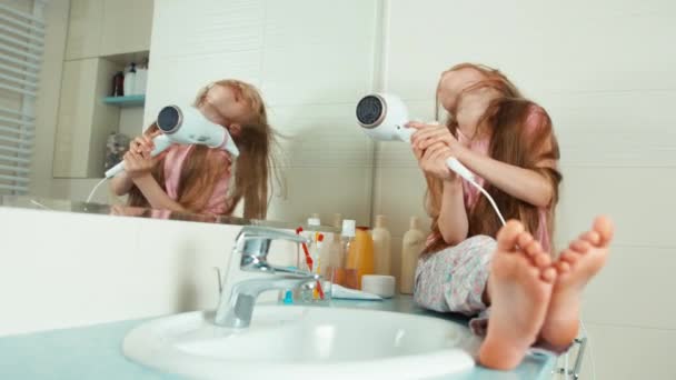 Chica de 7-8 años seca su largo cabello rubio hermoso con un secador de pelo en el baño y sonriendo a la cámara — Vídeos de Stock