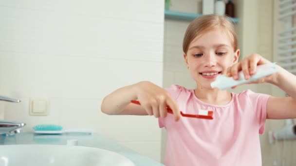 Muchacha retrato de cerca 7-8 años de edad limpieza de dientes con pasta de dientes y sonriendo a la cámara. Niño pone pasta de dientes en el cepillo — Vídeo de stock