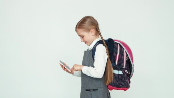 Schoolmeisje kind 7-8 jaar met rugzak met behulp van mobiele telefoon op witte achtergrond glimlachend op camera met tanden. Duim omhoog. OK — Stockvideo