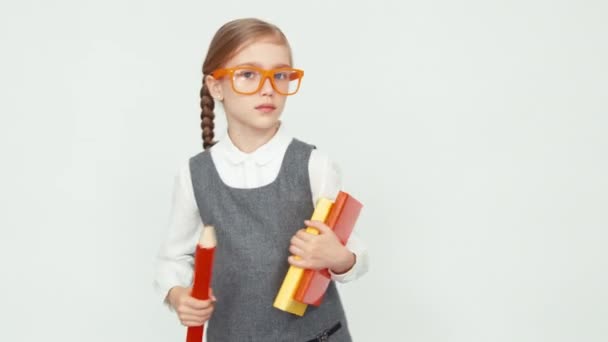 Μαθήτρια 7-8 ετών με γυαλιά κρατώντας βιβλία και ένα μεγάλο μολύβι. Απομονωμένα σε λευκό φόντο. Το παιδί μπαίνει στο πλαίσιο — Αρχείο Βίντεο
