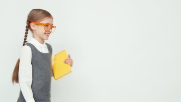Μαθήτρια 7-8 ετών χαμογελά στη φωτογραφική μηχανή. Κορίτσι με τα γυαλιά που κατέχουν ένα μεγάλο μολύβια και βιβλία. Παιδί εισέρχεται και εξέρχεται από το πλαίσιο σε λευκό φόντο — Αρχείο Βίντεο