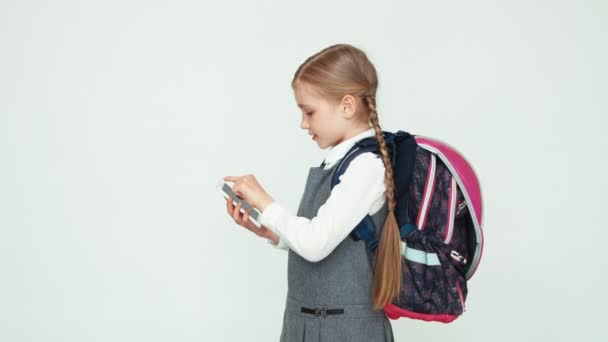 Портретна школярка 7-8 років з рюкзаком, використовуючи мобільний телефон на білому тлі, посміхаючись на камеру зубами — стокове відео