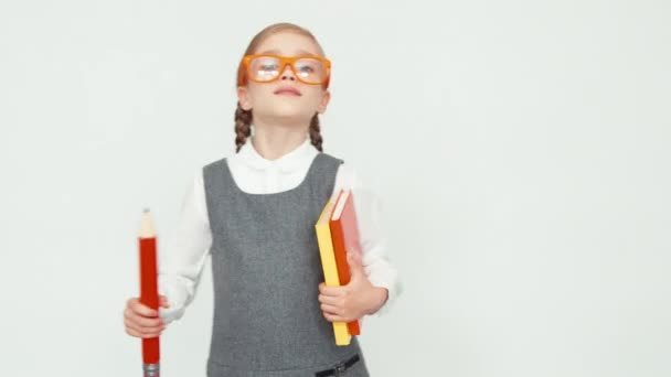 Школярка 7-8 років посміхається на камеру. Дівчина в окулярах тримає книжки і великий олівець. Дитина входить в рамку на білому тлі — стокове відео