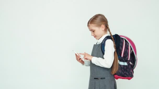 Skolflicka barn 7-8 år med ryggsäck med mobiltelefon på vit bakgrund tittar på kameran — Stockvideo