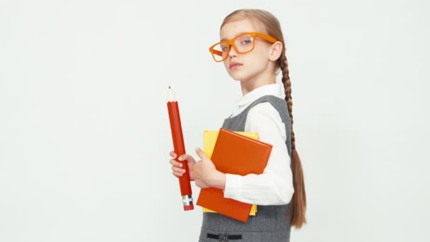 Colegiala niño de 7-8 años con gafas sosteniendo libros y un lápiz grande. Aislado sobre fondo blanco. Chica mirando a la cámara y asiente con la cabeza — Vídeos de Stock