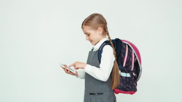 Colegiala niño 7-8 años con mochila usando teléfono móvil sobre fondo blanco sonriendo a la cámara con dientes — Vídeos de Stock