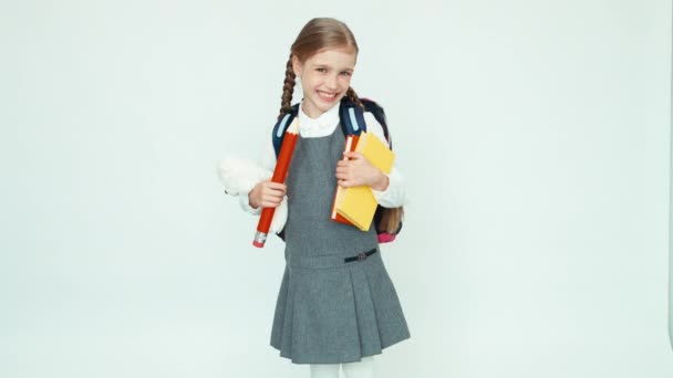 Carino scolaretta 7-8 anni con zaino in possesso di libri e grande matita e orsacchiotto e sorridente alla fotocamera su sfondo bianco — Video Stock