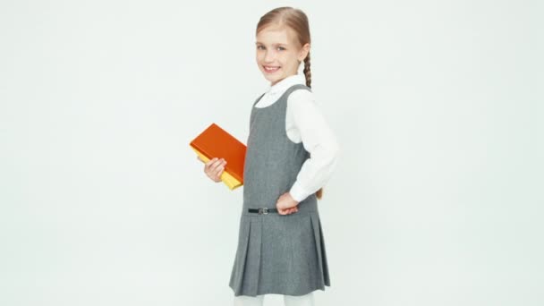 Portre sevimli kız öğrenci 7-8 yıl kitap tutan beyaz arka plan ve kamera dişli gülümseyen kamera arkasını döner. Yukarı başparmak. Tamam — Stok video