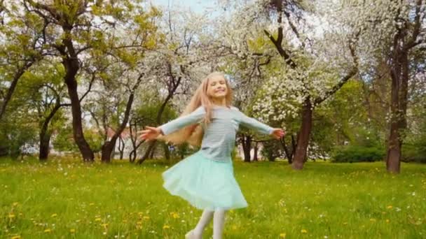 Söt flicka barn 7-8 år med blont långt hår spinning i parken under våren nära en blommande träd. Slow motion från Sony A6300 — Stockvideo