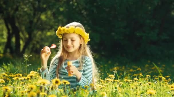 Retrato linda chica 7-8 años soplando burbujas de jabón y sentado en la hierba en el claro de dientes de león — Vídeo de stock