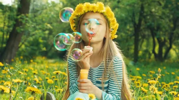 Portrait girl 7-8 years blowing soap bubbles and sitting in the grass on the glade of dandelions — Stock Video