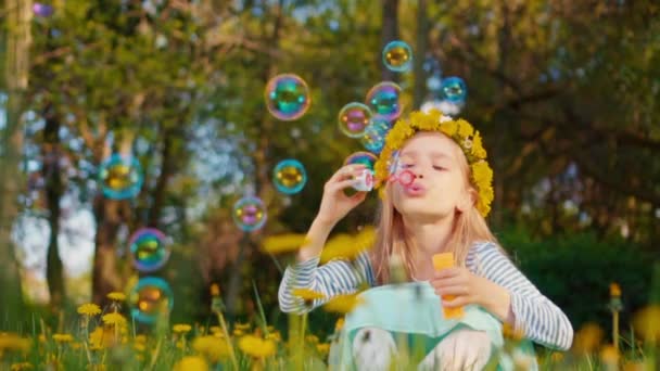 Portrét roztomilá dívka 7-8 let starý, posezení v trávě a usmívá a foukání mýdlové bubliny v parku. Zpomalený pohyb od Sony A6300 — Stock video