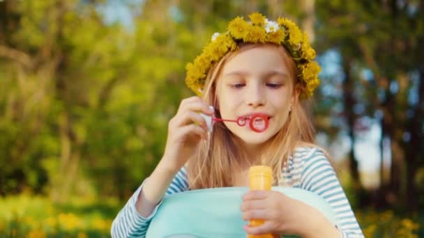 Zbliżenie portret dziewczyny 7-8 lat siedzi w trawie i uśmiechając się i dmuchanie baniek mydlanych w parku. Slow motion z Sony A6300 — Wideo stockowe