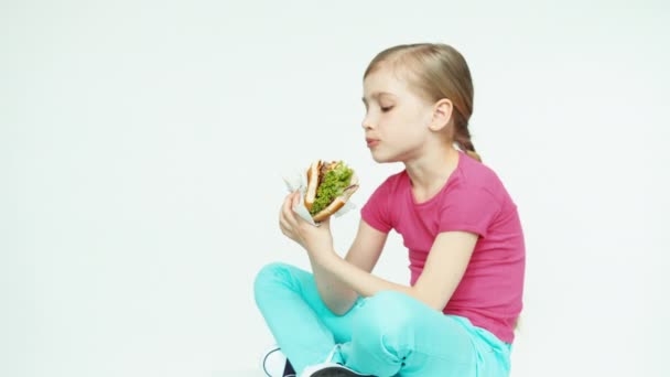 Flickor 7-8 år äter hamburgare och skratta och sitter på den vita bakgrunden. Reglaget — Stockvideo