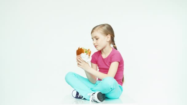 Ragazza bambino 7-8 anni guardando hamburger e mangiare hamburger e ridere e seduto sullo sfondo bianco. Pollice in alto. Va bene. — Video Stock