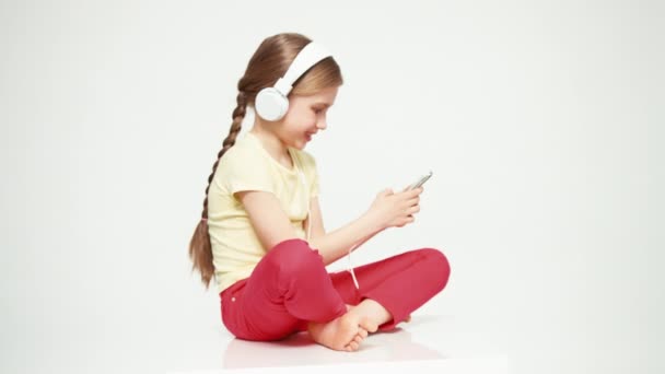 Дівчина 7-8 років використовує мобільний телефон і слухає музику на навушниках і сміється на камеру — стокове відео