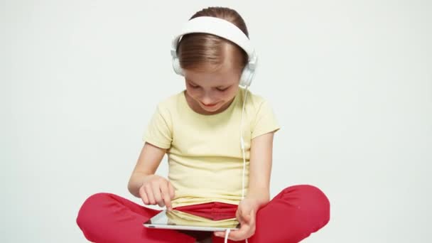 Портрет смеющейся девушки 7-8 лет с помощью планшетного ПК на белом фоне и прослушивания музыки на наушниках. Слайдер вышел — стоковое видео