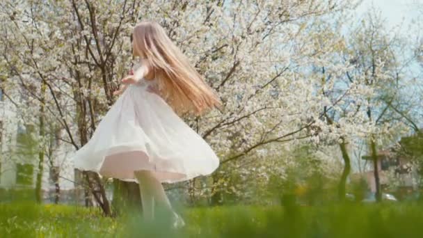 Chica rubia riendo girando en un vestido blanco sobre un fondo de árboles florecientes. Sony A6300 de cámara lenta — Vídeo de stock