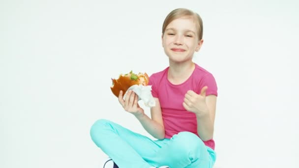 Closeup portret meisje kind 7-8 jaar oud kijken hamburger hamburger eten en lachen om de camera op de witte achtergrond. Duim omhoog. OK — Stockvideo