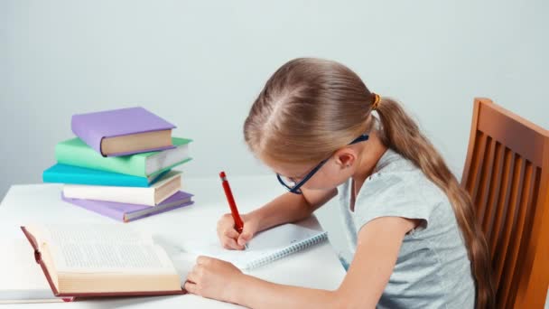 Retrato estudiante niña 7-8 años de edad en gafas algo escrito en su cuaderno y mirando a la cámara con sonrisa. El niño se rasguñó la nariz. Vista superior — Vídeo de stock