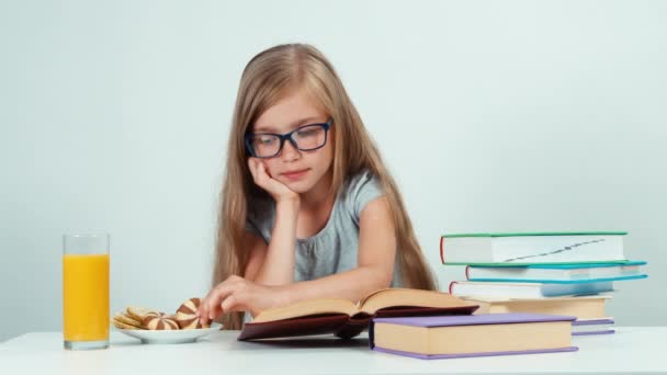 Retrato menina feliz 7-8 anos de idade em óculos lendo livro didático sobre a mesa e comer biscoito — Vídeo de Stock