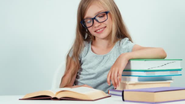 Colegiala retrato de cuatro ojos 7-8 años leyendo libro en la mesa sobre fondo blanco. Niño sonriendo a la cámara — Vídeos de Stock