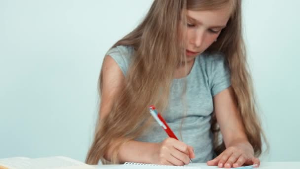 Cerrar retrato colegiala algo escrito en su cuaderno de la escuela sobre fondo blanco. Niña sentada en el escritorio. Panorama — Vídeo de stock