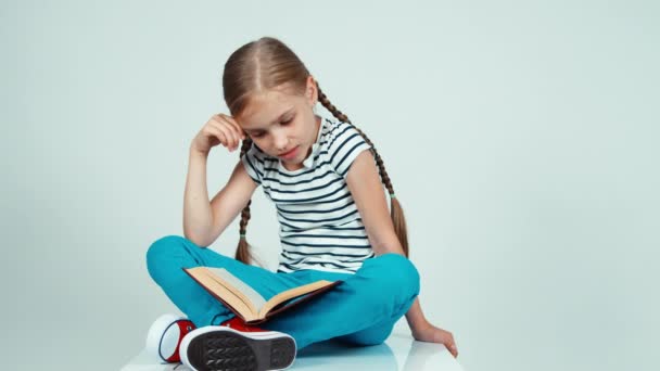 Портретная девочка читает книгу, сидя на полу и улыбаясь в камеру — стоковое видео