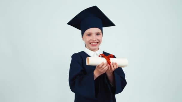 Портрет счастливая студентка выпускница девушка 7-8 лет в мантии холдинг и радуется ее диплом. Ребенок улыбается зубами на белом фоне — стоковое видео