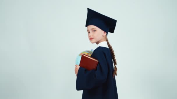 Portrait graduate girl 7-8 years in the mantle and hat turns at camera with globe and book on white background and laughing — Stockvideo