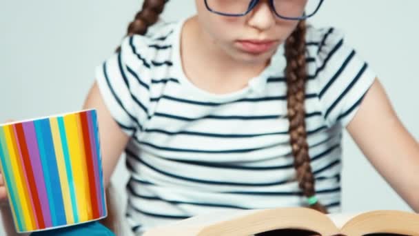 Close up retrato menina 7-8 anos de idade leitura livro e segurando xícara de chá isolado no branco. Panning — Vídeo de Stock