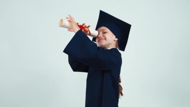 Retrato menina estudante feliz pós-graduação 7-8 anos no manto se alegra e se entrega com seu diploma e sorrindo com os dentes no fundo branco — Vídeo de Stock