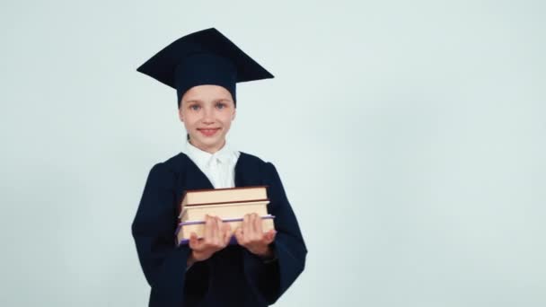 Graduate student girl in the mantle and hat turns around at camera with books on white background and smiling at camera with teeth. — Stock video