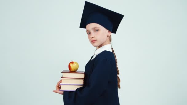 Primer plano retrato chica graduada 7-8 años en el manto y sombrero sosteniendo libros y manzana en blanco y sonriendo a la cámara con dientes. Niño gira en la cámara — Vídeos de Stock