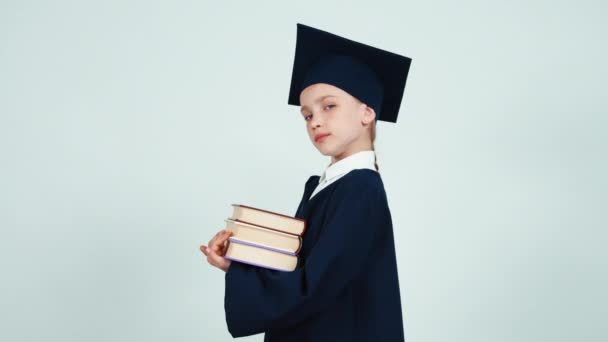 Graduada estudiante chica en el manto y sombrero sosteniendo sus libros sobre fondo blanco y sonriendo a la cámara con los dientes — Vídeo de stock