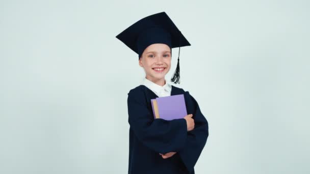 Laureata studente ragazza 7-8 anni nel mantello e cappello tenendo libro su sfondo bianco e sorridente alla macchina fotografica — Video Stock