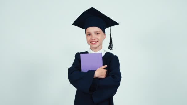 Yüksek lisans öğrencisi kız 7-8 yıl içinde beyaz arka plan ve kameraya gülümseyen kitap vermek manto ve şapka — Stok video