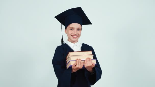 Graduada estudiante niña 7-8 años en el manto y sombrero sosteniendo sus libros sobre fondo blanco y sonriendo a la cámara con los dientes — Vídeo de stock
