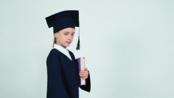 Portrait graduate girl 7-8 years in the mantle and hat entering at frame with book on white — Stockvideo