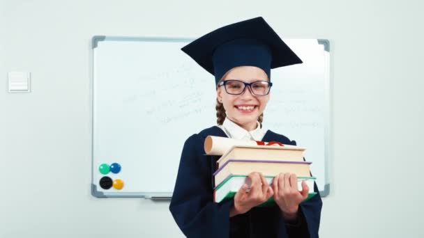 Student 7-8 jaar afgestudeerd in de mantel holding boeken en diploma schakelt op camera en lachen met tanden in de buurt van whiteboard. Schuifregelaar — Stockvideo