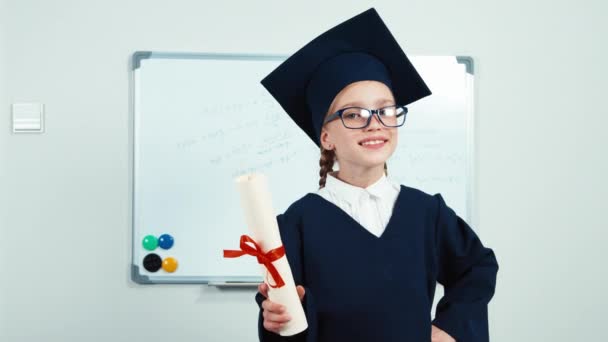 Estudante de retrato close-up 7-8 anos de pós-graduação no manto e chapéu segurando diploma e rindo com dentes perto whiteboard. Slider — Vídeo de Stock