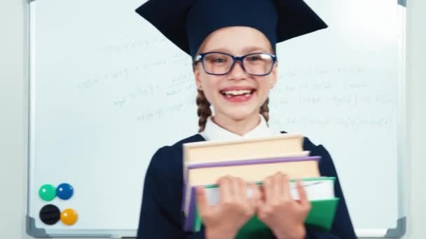 Estudiante de retrato de cerca 7-8 años se gradúa en el manto sosteniendo libros y diploma y riendo con dientes cerca de pizarra. Pulgar hacia arriba. Bien, control deslizante. — Vídeo de stock