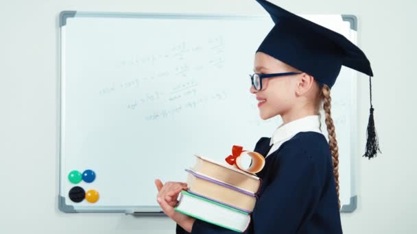 Gros plan portrait étudiant 7-8 ans diplômé dans le manteau tenant des livres et diplôme tourne à la caméra et rire avec des dents près du tableau blanc. Curseur — Video