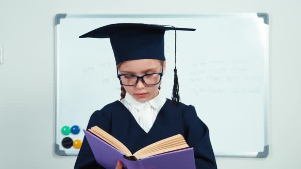 Close-up retrato menina estudante feliz 7-8 anos de pós-graduação no manto e chapéu lendo seu livro e sorrindo para a câmera com dentes perto whiteboard. Slider — Vídeo de Stock